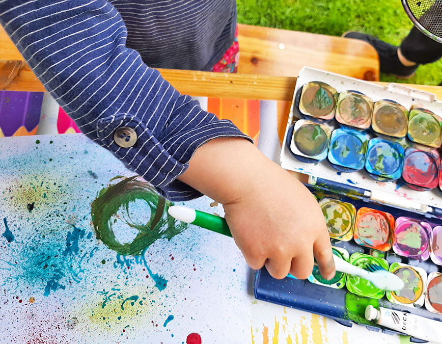 Hand eines kleinen Kindes mit Zahnbürste über buntem Farbmalkasten